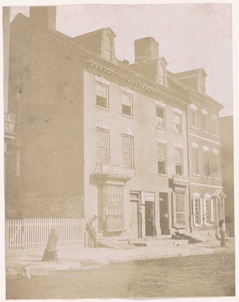 Salted paper photograph of a three-storey brick building in Philadelphia occupied by shops, identified by the Library of Congress as the first United States Mint